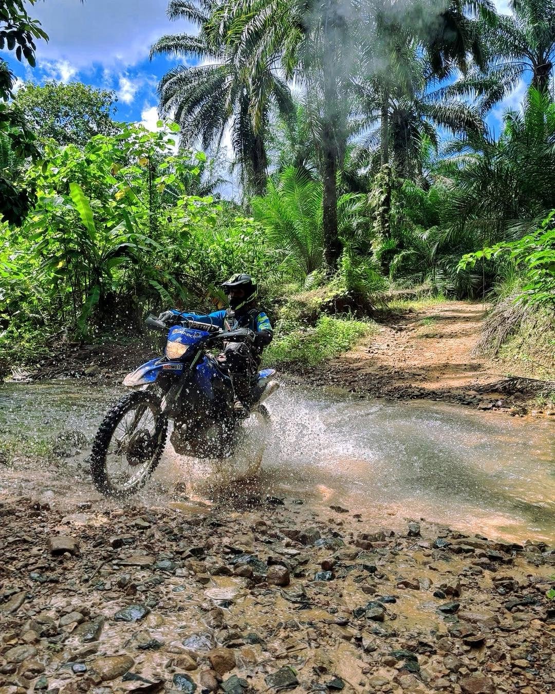 Enduro rider on a blue dirt bike splashing through a jungle stream in Krabi