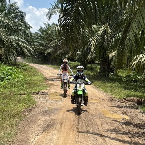 Dirt bike riders cruising along a flat dirt road through tropical palm trees in Krabi